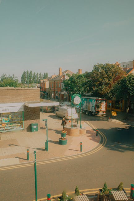 The image depicts a small urban commercial area on a sunny day, featuring a paved street with a sidewalk that includes a shopfront on the left side. The shopfront has a large window displaying various items and a canopy with signage. In front of the shop, there are decorative planter pots with small greenery and several green bollards serving as barriers to prevent vehicle encroachment. A pedestrian, dressed in dark clothing, is walking across the pedestrian crossing towards the shop. To the background, there are several parked vehicles, including a white car and a delivery truck with colorful branding parked along the street. Behind the vehicles, a row of residential buildings with brick facades and pitched roofs, some with chimneys, are visible amidst lush green trees that provide shade and scenic contrast. The environment appears clean and well-maintained, with clear weather casting natural light across the scene, illustrating a typical small-town commercial and residential streetscape suitable for local rubbish removal or private waste management services, such as those offered by Rubbish Clearance Barnet, to support on-site clearance or alternative waste handling in urban settings.
