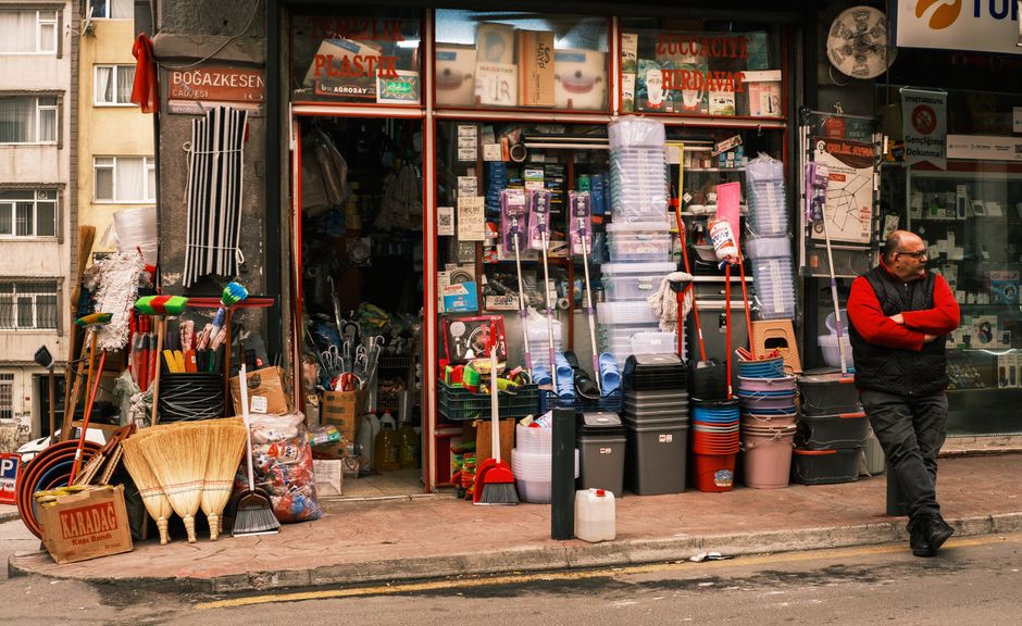 The image depicts a small urban commercial area on a sunny day, featuring a paved street with a sidewalk that includes a shopfront on the left side. The shopfront has a large window displaying various items and a canopy with signage. In front of the shop, there are decorative planter pots with small greenery and several green bollards serving as barriers to prevent vehicle encroachment. A pedestrian, dressed in dark clothing, is walking across the pedestrian crossing towards the shop. To the background, there are several parked vehicles, including a white car and a delivery truck with colorful branding parked along the street. Behind the vehicles, a row of residential buildings with brick facades and pitched roofs, some with chimneys, are visible amidst lush green trees that provide shade and scenic contrast. The environment appears clean and well-maintained, with clear weather casting natural light across the scene, illustrating a typical small-town commercial and residential streetscape suitable for local rubbish removal or private waste management services, such as those offered by Rubbish Clearance Barnet, to support on-site clearance or alternative waste handling in urban settings.