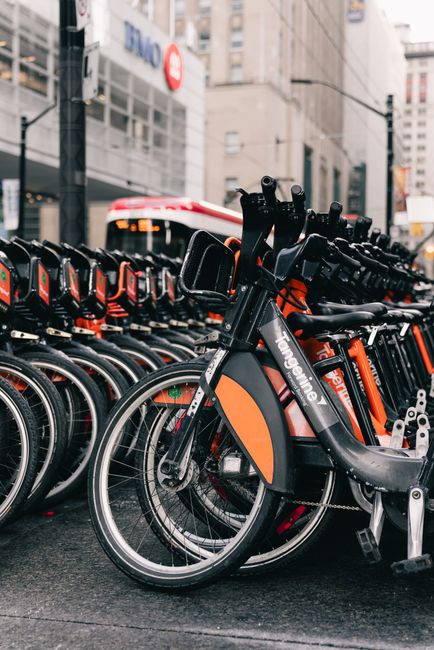 A row of black and orange rental bicycles parked in a designated bike-sharing station on a city street, with multiple bikes aligned closely together, some partially overlapping others. The bicycles have sturdy frames, textured black seats, and large wheels with silver spokes. The foreground bike's front wheel is turned slightly to the right, and the station stands on a paved surface. In the background, a modern urban environment is visible, including tall buildings with windows, commercial signage, and traffic signals. The scene appears well-lit, reflecting daytime natural light, and the overall environment suggests a busy city area suitable for alternative urban mobility options, such as bike rentals. The image emphasizes transportation infrastructure and aligns with topics related to sustainable mobility and city-based bicycle sharing schemes, similar to those operated by services like those seen in major cities and possibly managed by companies like Rubbish Clearance Barnet offering eco-friendly urban transport alternatives.