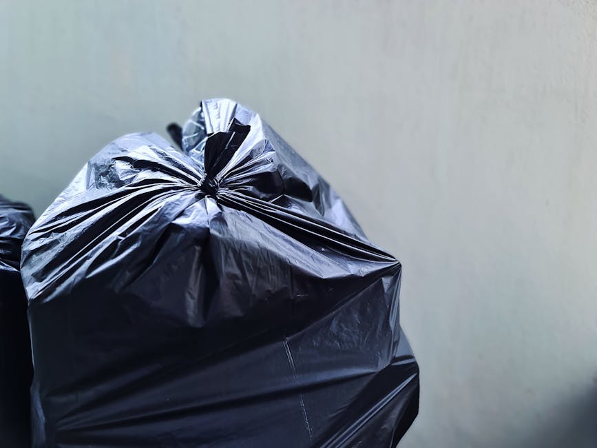 Close-up of a tightly knotted black plastic rubbish bag with a glossy surface, showing visible creases and slight tears in the material. The bag is positioned in the foreground, occupying the left half of the frame, with its top gathered and secured with a knot. In the background, other similar black plastic bags are stacked or leaning against each other, partially visible on the left side. The environment appears to be an indoor or covered area with a plain, light-colored wall behind the bags, providing minimal environmental detail. The lighting is diffuse, casting soft shadows and giving a neutral tone to the scene, typical of an area prepared for rubbish collection or disposal. This image exemplifies a common scene in private waste handling, often associated with rubbish clearance services such as those offered by Rubbish Clearance Barnet, which specializes in alternative waste removal options outside of local authority collection systems.