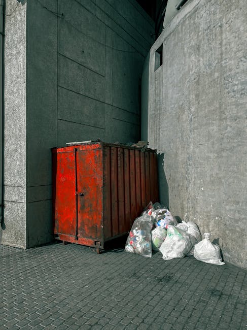 A large collection of overflowing rubbish bins and scattered waste bags positioned on a paved sidewalk in front of a commercial building with storefronts, including a fish bar. The waste includes various types of rubbish such as black and red tipped garbage bags, flattened cardboard boxes, crumpled paper, plastic packaging, and miscellaneous discarded items. Several bins are visible, including a grey mixed paper and cardboard bin with a partially open lid, a black bin, and a red recycling bin, all filled beyond capacity, causing waste to spill onto the ground. The background features a building under construction or renovation with scaffolding and safety fencing, and a parked silver car is seen on the left side behind a metal railing. The scene is lit with natural daylight, and the environment suggests a busy town center or shopping precinct area, where private waste collection or rubbish clearance services might be needed to manage on-site waste handling.