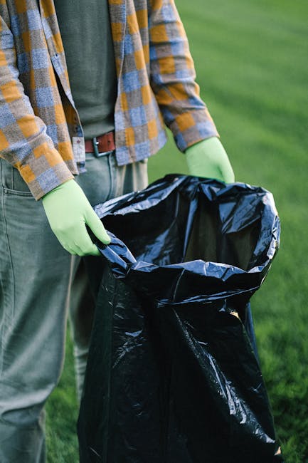 A person, dressed in a plaid shirt with yellow and blue tones over a green T-shirt, wearing grey trousers secured with a brown belt, is holding open a large black rubbish bag lined with a shiny silver interior. The person is wearing light green gloves and is standing outdoors on a grassy area with lush green grass visible in the background. The rubbish bag appears to be intended for garden or household waste collection, and the scene suggests preparation for rubbish removal or waste disposal, consistent with private waste handling services offered by companies like Rubbish Clearance Barnet. The natural lighting and outdoor setting emphasize the practical nature of rubbish collection and disposal work, supporting the context of environmental management and rubbish removal services as part of site clearance or waste collection activities.


