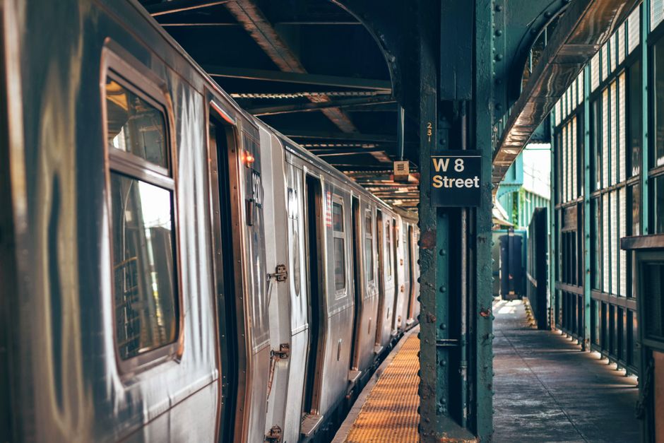 A silver subway train is stationary at a platform under a metal structure on a city street, with reflections visible on the train’s metallic surface. The train doors are closed, and the windows reveal glimpses of the interior. The platform features a textured yellow safety strip along the edge, with a black metal post labeled 'W 8 Street' positioned beside it. To the right, a row of glass windows and metal supports run along the length of the platform, allowing natural light to illuminate the scene. The platform floor appears concrete, with some debris and shadows cast by the overhead structure. The environment suggests an urban setting, with the station's steel framework and enclosed design providing a semi-private space for passengers. This scene is a typical view of city transit infrastructure, often associated with public transportation or alternative approaches to urban travel, connecting well with concepts of independent travel and space management in city environments, relevant to the broad scope of rubbish removal services like those offered by Rubbish Clearance Barnet.